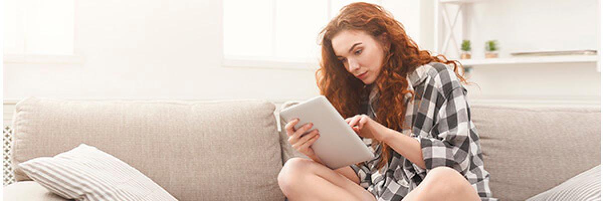 Cuenta de Pago Básica - Chica joven con una camisa de cuadros sentada en el sofá de su casa con una tablet buscando cuentas